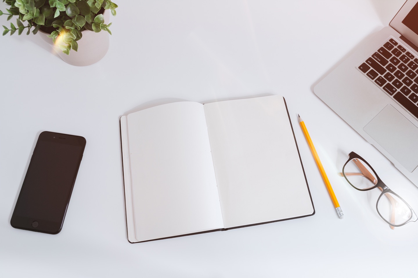 An overhead picture of a white desk with a laptop, smart phone, pair of glasses, pencil and blank notebook laid out on it. 