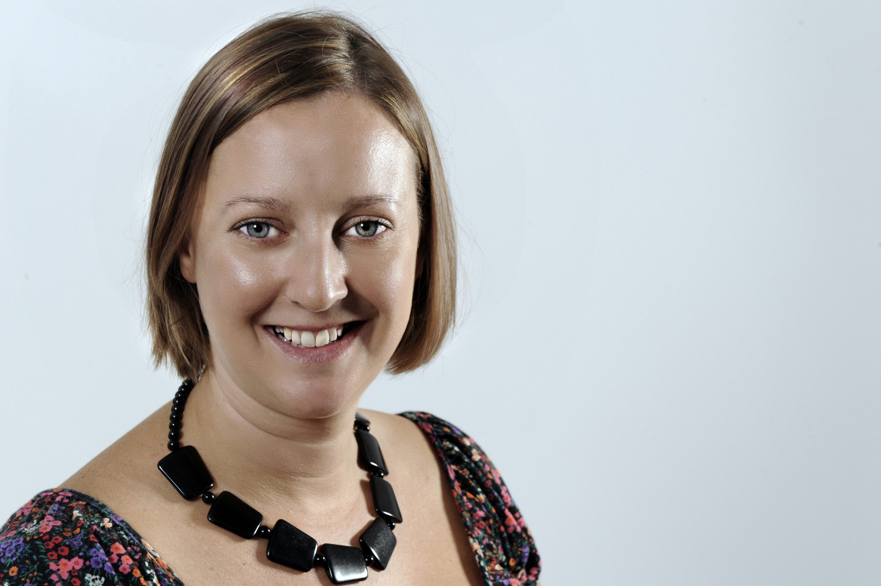 Photo of a white woman with a mousy brown straight bob. It is a professional head shot with her smiling at the camera, wearing a chunky black necklace and flowery t-shirt. The background is a light grey/blue blank studio.  