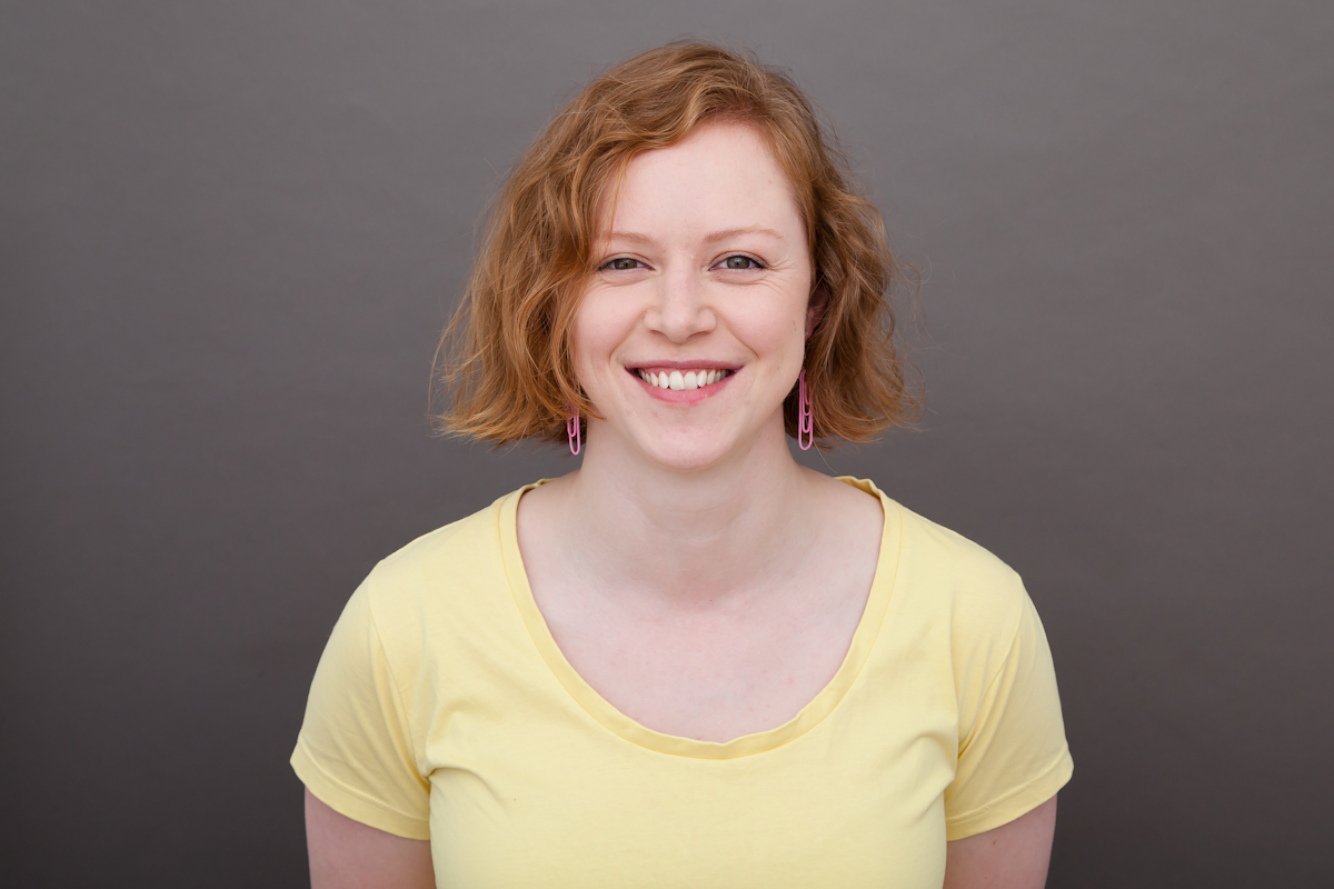 Katie Goudie is wearing a yellow tshirt and pink paperclip earrings. She is standing in front of a grey backdrop and is smiling widely.